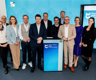 Gruppenfoto von elf Personen, die vor einem blauen Hintergrund mit dem Logo 'Bayern-Allianz gegen Desinformation' stehen. Die Gruppe besteht aus Männern und Frauen in formeller und halbformeller Kleidung, die sich um ein Rednerpult versammelt haben.