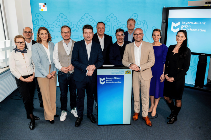 Gruppenfoto von elf Personen, die vor einem blauen Hintergrund mit dem Logo 'Bayern-Allianz gegen Desinformation' stehen. Die Gruppe besteht aus Männern und Frauen in formeller und halbformeller Kleidung, die sich um ein Rednerpult versammelt haben.