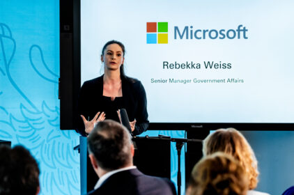 Eine Frau in einem schwarzen Anzug und schwarzem Pullover steht an einem Rednerpult vor einem großen Bildschirm, der das Microsoft-Logo und den Text 'Rebekka Weiss, Senior Manager Government Affairs' anzeigt. Sie spricht zu einem Publikum vor einem hellblauen Hintergrund mit abstrakten Mustern.