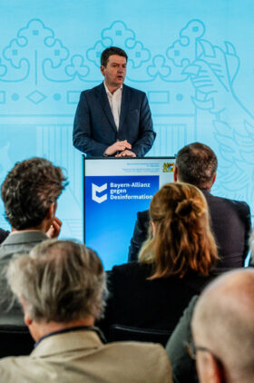 Ein Mann in einem dunkelblauen Anzug steht an einem Rednerpult mit dem Logo 'Bayern-Allianz gegen Desinformation'. Er spricht zu einem Publikum, das von hinten zu sehen ist, vor einem hellblauen Hintergrund mit abstrakten Mustern.