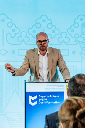 Ein Mann in einem beigen Jackett und weißen Hemd steht an einem Rednerpult mit dem Logo 'Bayern-Allianz gegen Desinformation'. Er spricht zu einem Publikum, mit einer Geste der rechten Hand, vor einem hellblauen Hintergrund mit abstrakten Mustern.